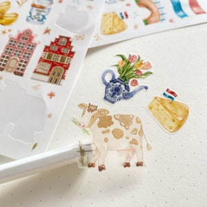 May include: A set of colorful stickers featuring Dutch-themed illustrations. The stickers include a cow, a house, a teapot with flowers, and a wedge of cheese.