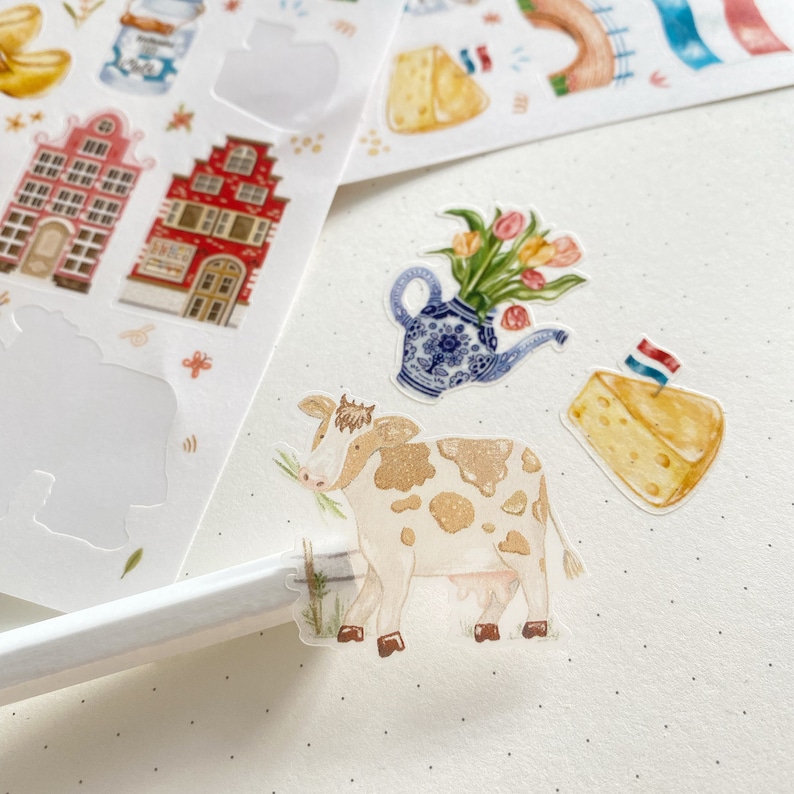 May include: A set of colorful stickers featuring Dutch-themed illustrations. The stickers include a cow, a house, a teapot with flowers, and a wedge of cheese.