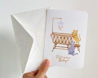May include: A white greeting card with a watercolor illustration of a baby cradle, a mobile with a cloud, moon, and stars, and two cats. One orange cat stands by the cradle, and a gray cat sits below. The text Welcome Baby is printed below.