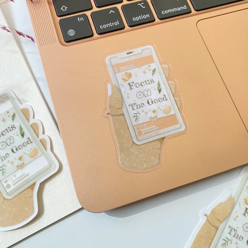 Puede incluir: Una pegatina de una mano sosteniendo un tel&eacute;fono con el texto "Focus on the Good" en la pantalla. El tel&eacute;fono tiene un dise&ntilde;o floral y un fondo marr&oacute;n claro.