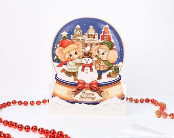 May include: A die-cut greeting card shaped like a snow globe featuring two cartoon characters building a snowman in front of European-style buildings. The base has a red ribbon and the text Happy Holidays printed in brown script lettering.