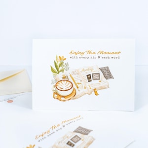 Peut inclure: Carte de vœux blanche avec une illustration à l'aquarelle d'une tasse de café, d'un livre ouvert et d'éléments floraux. La carte porte l'inscription "Enjoy The Moment with every sip & each word" en lettres dorées. Une enveloppe assortie est visible.