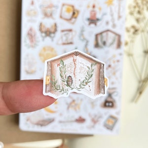 May include: A sticker of an open book with brown pages and green leaves. The book is open to a page with handwritten text and a small illustration of a flower.