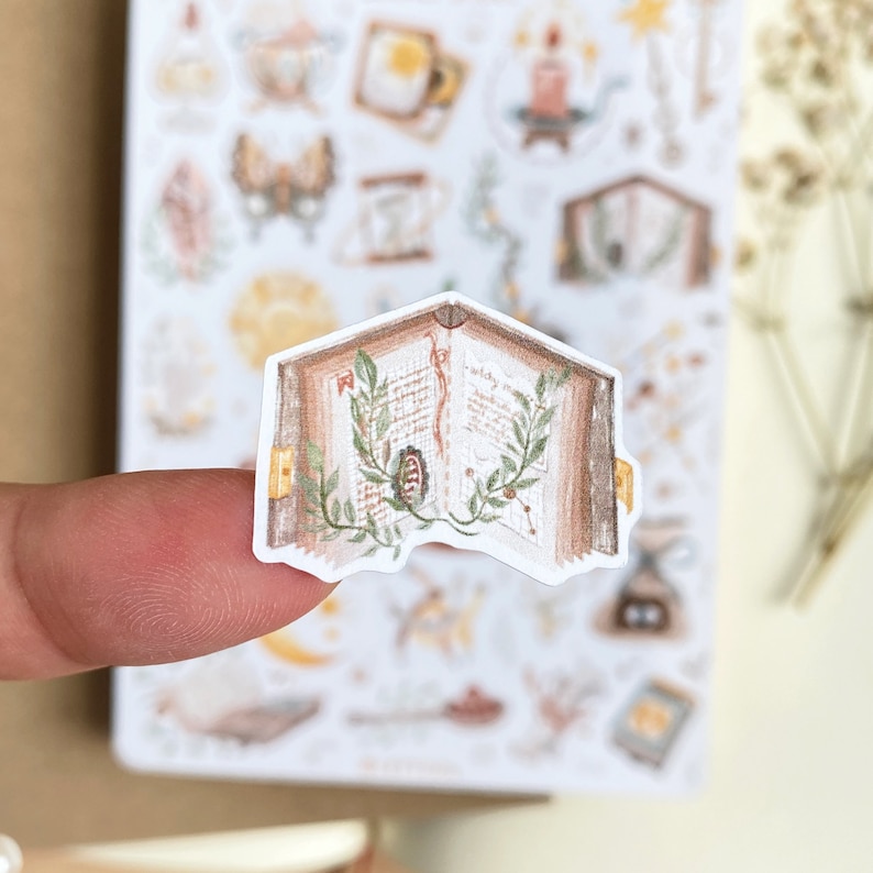 May include: A sticker of an open book with brown pages and green leaves. The book is open to a page with handwritten text and a small illustration of a flower.