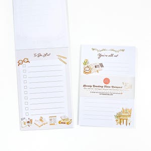 Puede incluir: Dos blocs de notas blancos con ilustraciones decorativas. Uno dice "To Do List" con gafas y un lápiz. El otro dice "You're all set" y "Lovely Reading Time Notepad". Los blocs de notas miden 15 x 10 cm.