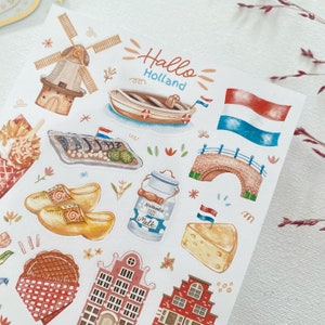May include: A sheet of stickers featuring illustrations of Dutch culture, including a windmill, clogs, a canal bridge, a boat, cheese, and a milk jug. The stickers are colorful and whimsical, with a handwritten title that reads "Hallo Holland".