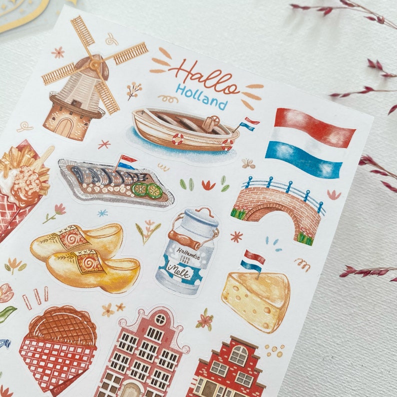 May include: A sheet of stickers featuring illustrations of Dutch culture, including a windmill, clogs, a canal bridge, a boat, cheese, and a milk jug. The stickers are colorful and whimsical, with a handwritten title that reads "Hallo Holland".