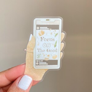 Puede incluir: Una pegatina transparente para tel&eacute;fono con una mano sosteniendo un tel&eacute;fono. La pantalla del tel&eacute;fono muestra el texto "Focus on The Good" con ilustraciones florales.