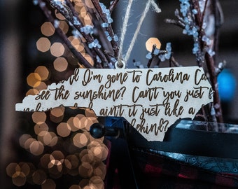 May include: A wooden North Carolina-shaped ornament with the text "and I'm gone to Carolina Can't you just see the sunshine?" The ornament is suspended by a string, set against a blurred backdrop of lights and branches.