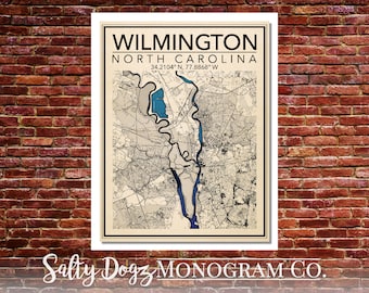 Puede incluir: Una impresión de mapa de estilo vintage de Wilmington, Carolina del Norte, con un esquema de color azul y turquesa. El mapa muestra las calles, vías fluviales y puntos de referencia de la ciudad. El texto "WILMINGTON NORTH CAROLINA 34.2104° N, 77.8868° W" está impreso en la parte superior del mapa. El texto "Salty Dogz MONOGRAM CO." está impreso en la parte inferior del mapa.