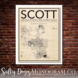 May include: A vintage-style map print of Scott Air Force Base in Saint Clair County, Illinois. The map shows the base layout and surrounding area. The print includes the base name, location, and coordinates: 38.4008° N, 89.9253° W. The print is framed in a white frame and has a beige background.