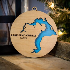 May include: A wooden Christmas ornament shaped like Lake Pend Oreille in Idaho. The lake is cut out of the wood and painted blue. The ornament is hanging from a string.
