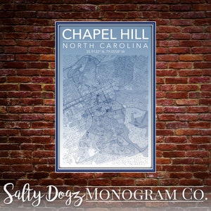 Op de afbeelding: Een blauwe en witte kaart van Chapel Hill, North Carolina, met de coördinaten 35.9132° N, 79.0558° W. De kaart is ingelijst en hangt aan een bakstenen muur. De tekst "Salty Dogz Monogram Co." staat onder de kaart.