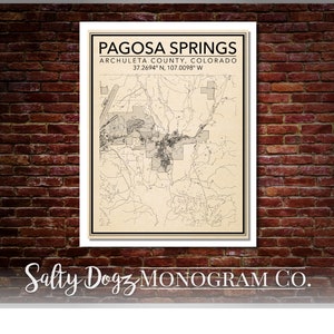 May include: A vintage-style map of Pagosa Springs, Archuleta County, Colorado. The map is printed on a cream-colored background with black lines and text. The text includes the name of the town, the county, and the coordinates 37.2694° N, 107.0098° W. The map is framed in a white frame.