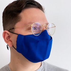 Op de afbeelding: Een persoon draagt ​​een blauw mondkapje en een ronde, goudkleurige bril. De persoon draagt ​​een grijs T-shirt.