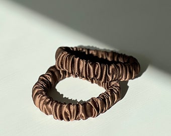 May include: Two brown silk scrunchies are displayed on a white surface. The scrunchies are made of a soft, gathered fabric, and appear to be new. The lighting creates shadows that highlight the texture and shape of the hair accessories.