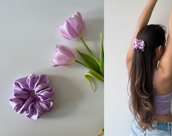 May include: Two purple satin scrunchies, one on a white background and one in a person's hair. The scrunchies are made of soft, silky fabric and have a gathered, ruffled look.