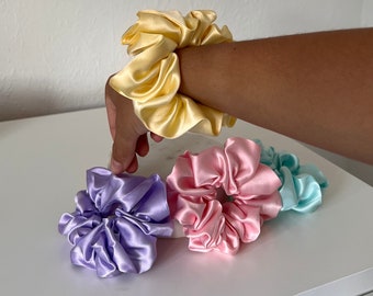 May include: A collection of satin scrunchies in various colors, including yellow, lavender, pink, and teal. The scrunchies are made of a shiny, smooth fabric and are gathered to create a voluminous look. One yellow scrunchie is on a wrist.