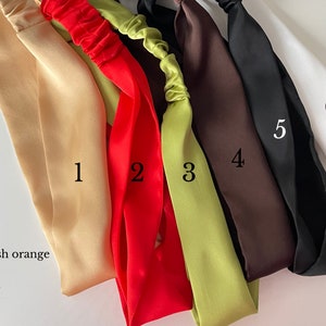 May include: Six satin headbands in different colors: gold, reddish orange, green, brown, black, and white. The headbands are arranged in a row, with the gold headband on the left and the white headband on the right.