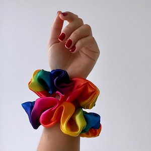 May include: Two rainbow-colored scrunchies made of satin fabric. The scrunchies are stacked on top of each other on a person's wrist.