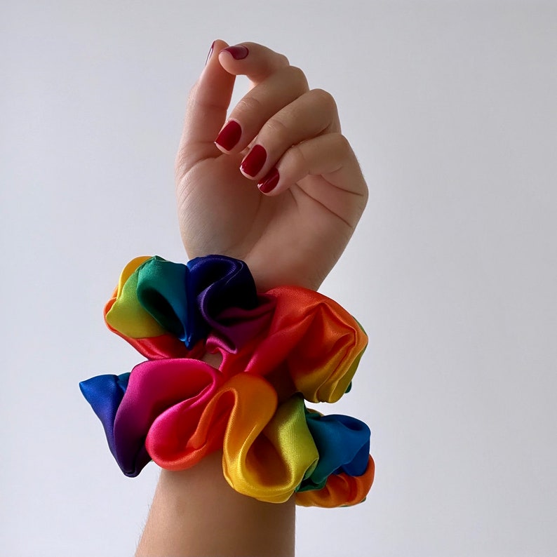 May include: Two rainbow-colored scrunchies made of satin fabric. The scrunchies are stacked on top of each other on a person's wrist.