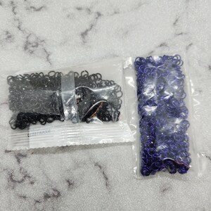 Op de afbeelding: Twee kleine plastic zakjes gevuld met metalen sprongringen. Eén zakje bevat zwarte sprongringen en de andere paarse. De sprongringen zijn klein, cirkelvormig en lijken van metaal te zijn.