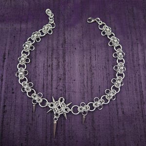 May include: A silver chainmail choker necklace with a central starburst design and dangling spikes. The necklace features a floral pattern along the chain and a lobster clasp closure. The jewelry is displayed on a purple background.