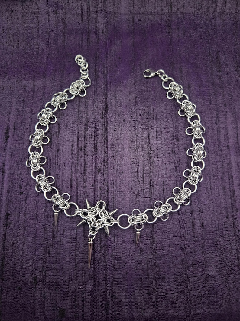May include: A silver chainmail choker necklace with a central starburst design and dangling spikes. The necklace features a floral pattern along the chain and a lobster clasp closure. The jewelry is displayed on a purple background.