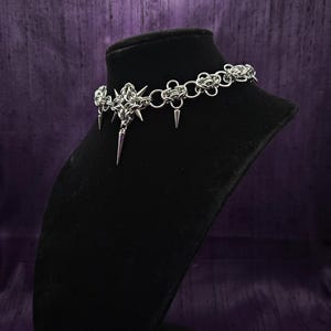 May include: A silver-colored choker necklace with a gothic design. The necklace features a central pendant with a starburst design and dangling spikes. The choker is displayed on a black mannequin head against a purple background.