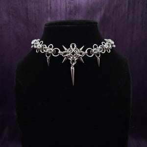May include: A silver choker necklace with a gothic design. The necklace features a central starburst pendant with dangling spikes and chainmail-style links. The necklace is displayed on a black mannequin head against a purple backdrop.