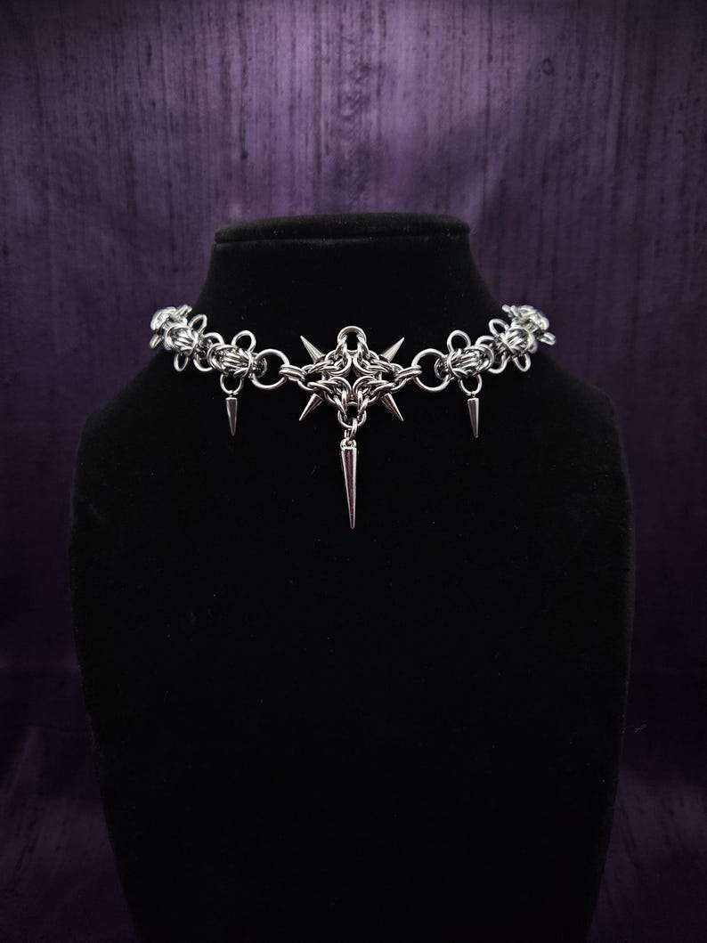 May include: A silver-colored choker necklace with a gothic design. The necklace features a central starburst pendant with dangling spikes and chainmail-style links. The necklace is displayed on a black mannequin head against a purple backdrop.
