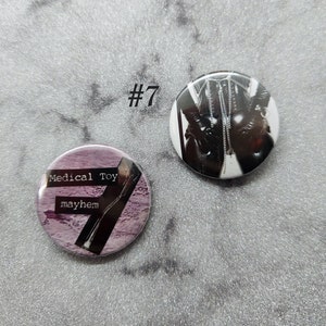 Könnte beinhalten: Zwei Anstecknadeln. Eine Nadel zeigt ein Schwarzweißbild einer Person in Lederkleidung. Die andere Nadel ist lila und trägt den Text "Medical Toy" und "mayhem".