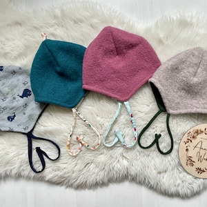 Könnte beinhalten: Vier bunte Baby-Mützen mit Ohrenklappen und Bindebändern, präsentiert auf einer flauschigen weißen Oberfläche. Die Mützen sind in Grautönen mit Wal-Print, Türkis, Mauve und Hellgrau gehalten. Jede Mütze hat ein andersfarbiges Band.