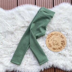 Könnte beinhalten: Salbeigrünes geripptes Leggings auf einem weißen, flauschigen Teppich. Ein rundes Holzschild mit dem Text "Anna's Workshop" und floralen Akzenten ist ebenfalls vorhanden. Die Leggings sind aus einem weichen, strukturierten Stoff gefertigt.