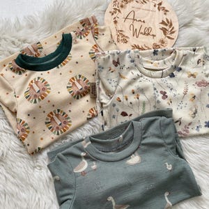 May include: Three children's t-shirts with different patterns. One is beige with lion faces, another is cream with floral and butterfly designs, and the third is teal with geese. A wooden plaque with text is also present.