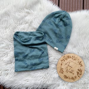 Könnte beinhalten: Ein tealfarbenes Mützen- und Halstuch-Set mit einem sich wiederholenden Traktormuster. Das Set wird auf einer flauschigen weißen Oberfläche präsentiert. Ein rundes Holzschild mit floralem Design und Text ist ebenfalls vorhanden.