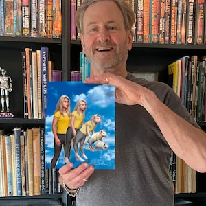 May include: A man is holding a book cover featuring two women and three white rats in a blue sky with white clouds. The book cover has the title "Infinite Worlds" and the author's name "Vincent Di Fave".