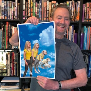 May include: A person holds up a print of a fantasy illustration. The illustration depicts two women and three anthropomorphic rats in a blue sky with white clouds. The women are wearing yellow shirts and grey pants. The rats are white and grey with yellow fur. The print is on white paper with a white border.