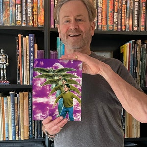 May include: A man holds a book cover featuring a purple sky with white clouds and three figures flying with wings. The book title is "Drawing the Head and Hands" and "Infinite Worlds" by Vincent Di Fate.