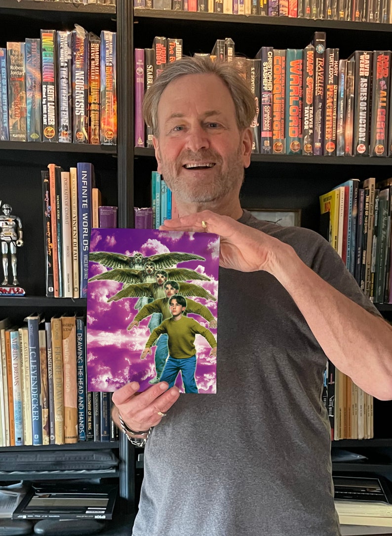 May include: A man holds a book cover featuring a purple sky with white clouds and three figures flying with wings. The book title is "Drawing the Head and Hands" and "Infinite Worlds" by Vincent Di Fate.