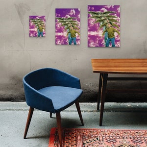 May include: Three framed prints of a person flying with wings against a purple and white cloudy background. The prints are different sizes and are hanging on a gray wall. A blue chair and a wooden table are in the foreground.