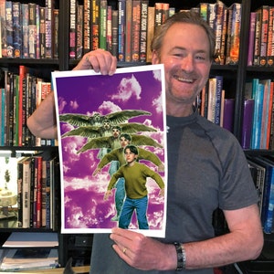 May include: A man holds up a print of a fantasy illustration. The illustration features three figures with wings, flying against a purple and white cloudy sky. The figures are wearing casual clothing and have a whimsical expression.