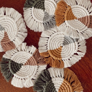 May include: A collection of handmade macrame coasters. Each coaster features a circular design with a fringed edge and a segmented color pattern in shades of white, brown, gray, and green. The coasters are arranged on a wooden surface.