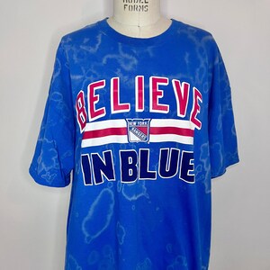 May include: A blue tie-dye t-shirt with the words "Believe in Blue" and the New York Rangers logo.