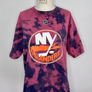 May include: A tie-dye t-shirt with a faded red and black color scheme. The shirt features a large orange and white New York Islanders logo with the text "NY ISLANDERS".