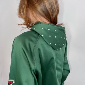 May include: A dark green cropped hoodie with a white rhinestone design on the hood. The hoodie has a red and white patch on the left sleeve.