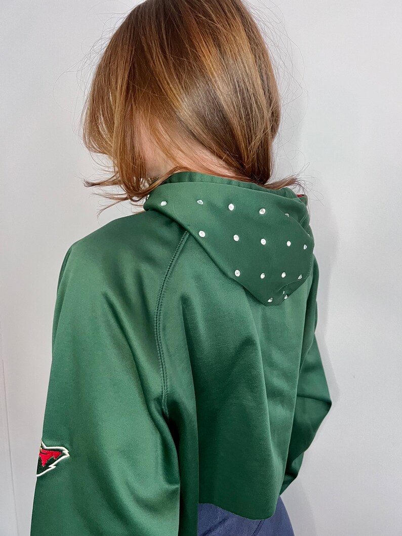 May include: A dark green cropped hoodie with a white rhinestone design on the hood. The hoodie has a red and white patch on the left sleeve.