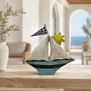 Pode incluir: Uma estatueta decorativa de veleiro com uma vela cinza-claro texturizada, um detalhe de sol amarelo brilhante e um casco azul com uma faixa preta e três pequenos pontos vermelhos. No topo, há uma bandeira triangular com uma cruz grega.