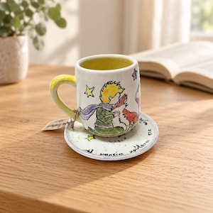 May include: A white ceramic mug and matching saucer featuring a hand-painted illustration of a young person with yellow hair and a red fox. The mug has a yellow handle and a yellow interior. The saucer is decorated with small black stars and text.