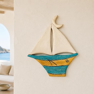 Puede incluir: Una escultura de pared de un velero de cerámica con dos velas triangulares de color blanco roto rematadas con una forma de media luna. El casco está decorado con rayas horizontales en tonos alternos de azul cerceta y amarillo mostaza.